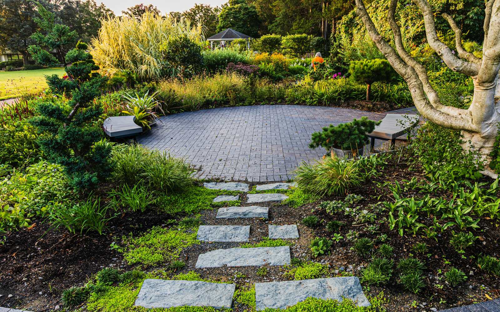 Stone stepping stones leading to a round block-paved patio with benches, framed by planted borders